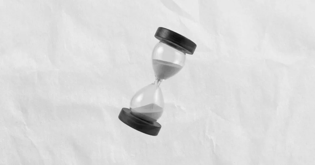 Reloj de arena minimalista inclinado con extremos de madera circular, mostrando la arena suspendida en el centro sobre un fondo de textura de papel arrugado.